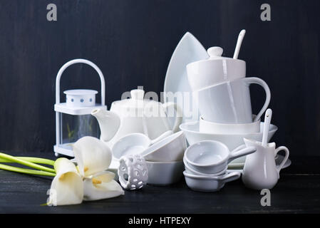 Set of new white dishes with teapot, tea cups, and plates on a black wooden table with fresh callas Stock Photo
