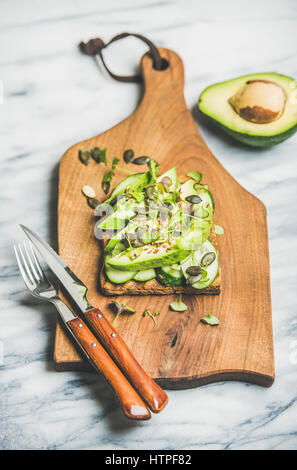 Healthy green veggie breakfast concept. Sandwich with avocado, cucumber, kale, kress sprout, pumpkin seeds on wooden board over grey marble background Stock Photo