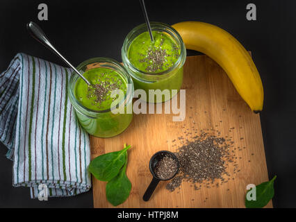 Green spinach, chia seed, banana and protein powder smoothie in two jars on the bamboo wooden surface, cloth napkin, dark background. Healthy breakfas Stock Photo