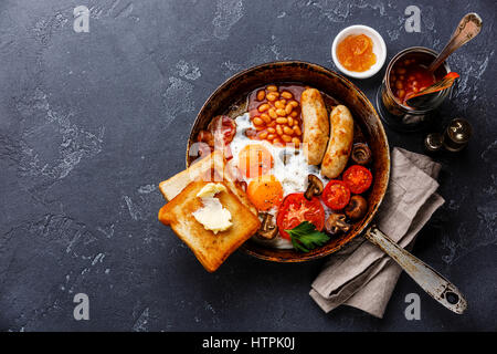 English Breakfast in cooking pan with fried eggs, sausages, bacon, beans and bread toasts on dark stone background copy space Stock Photo