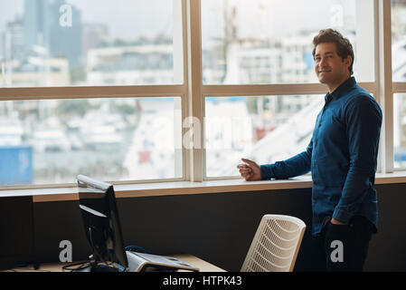 Portrait of a casually dressed young businessman smiling while standing in an office by windows overlooking a view of the city skyline Stock Photo