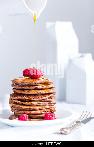 Stack of homemade american eggless chocolate pancakes with maple syrup flowing from jar, served on white plate  along with raspberries. Stock Photo