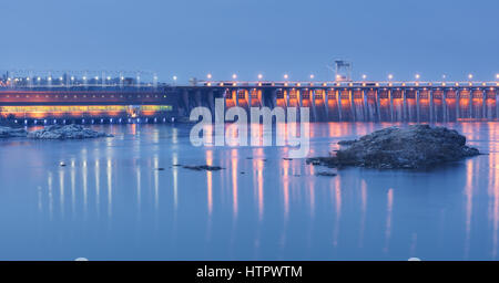 Dam at night. Beautiful industrial landscape with dam hydroelectric power station, bridge, river, city illumination reflected in water, rocks and sky. Stock Photo