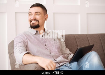 At the weekends. Positive joyful bearded man using a tablet and being in a great mood while resting at home Stock Photo