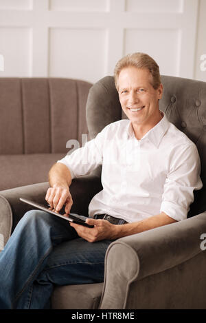 Modern technology. Nice cheerful good looking man sitting in the armchair and using a laptop while working at home Stock Photo