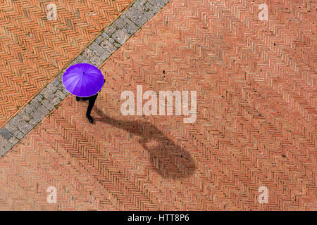 Anonymous man in black suit is walking on red and brown tiles square in Italy, hiding from the Sun with a purple umbrella. His shadow could be seen. Stock Photo