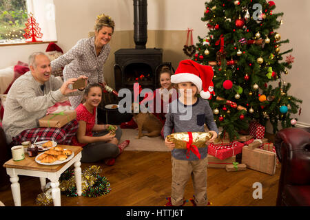 A family are sat by the Christmas Tree holding presents on christmas morning. Dad is taking a photo of his son holding a gift. Stock Photo
