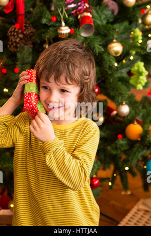 Little boy shaking a present close to his ear to try and guess what it is. He is standing in front of the Christmas tree. Stock Photo