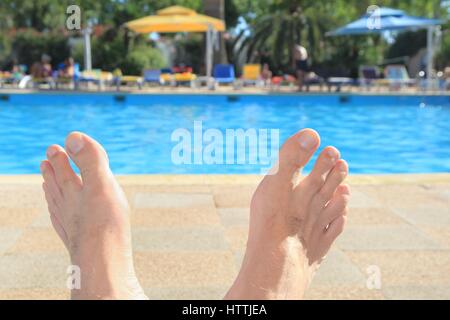 Man's feet in sunshine with swimming pool, umbrellas and sunloungers in the background Stock Photo