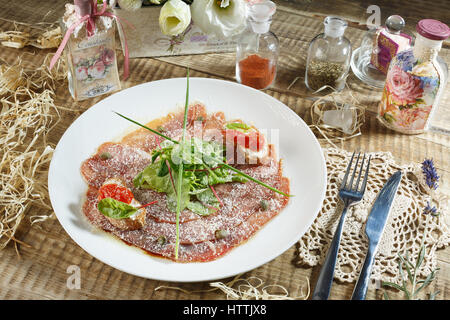 Sliced meat with spices, tomato cherry, bread, knife, fork plate on wooden background. Stock Photo