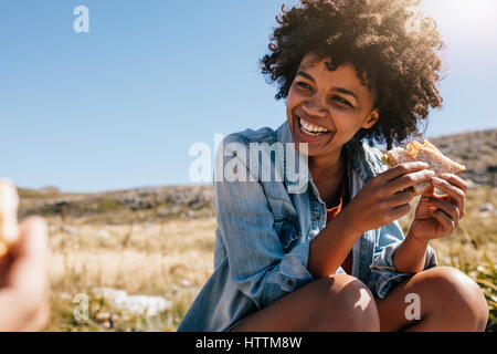 Happy young african woman eating sandwich and smiling. Taking break during country hike. Stock Photo
