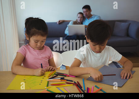 Boy and girl drawing in paper while parents using laptop in background Stock Photo
