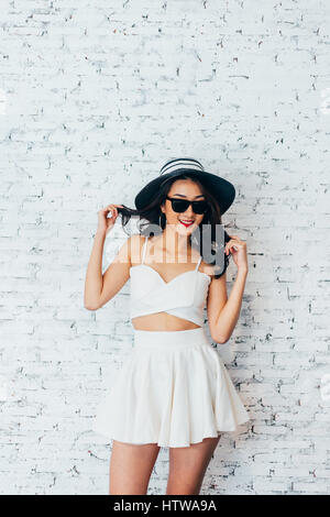 Young happy Asian woman smiling in fashionable dress and sunglasses and summer hat over white brick wall with copy space Stock Photo