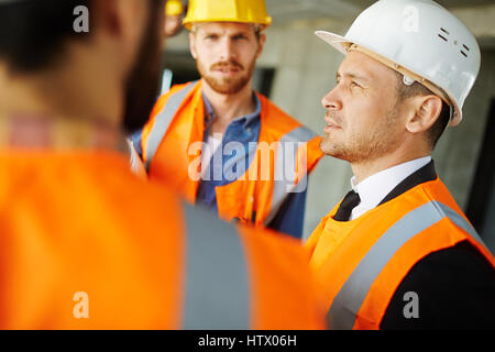 Confident builder in helmet and uniform talking to his colleagues Stock Photo