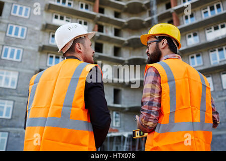 Backs of two builders in uniform having discussion on background of unfinished edifice Stock Photo