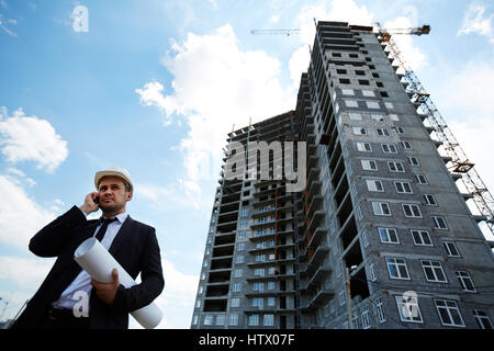 Elegant investor in helmet talking on mobile phone by unfinished building Stock Photo