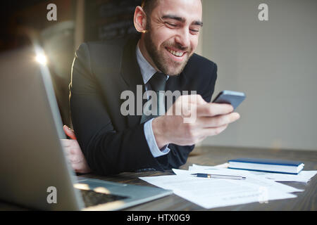 Portrait of modern bearded businessman wearing black formal suit working in office with documentation and laptop, smiling cheerfully while reading tex Stock Photo