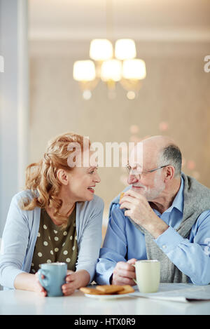 Portrait of smiling senior couple sitting close together at kitchen table with tea cups and homemade cookies, talking and laughing looking caringly at Stock Photo