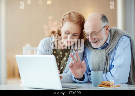 Portrait of beautiful elderly couple waving to laptop camera saying Hello video chatting with family using modern laptop and smiling happily Stock Photo