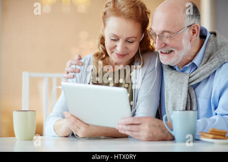 Portrait of smiling elderly couple using tablet watching videos with earphones looking at screen and embracing home at table Stock Photo
