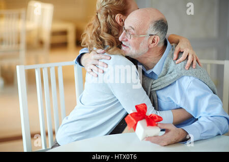 Portrait of loving mature couple embracing happily celebrating holiday together at home, woman hugging gray haired man holding gift box at table Stock Photo