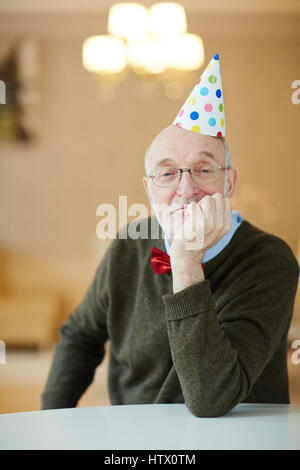 Portrait of nice old man wearing party hat sitting alone at table waiting for guests looking pensive Stock Photo