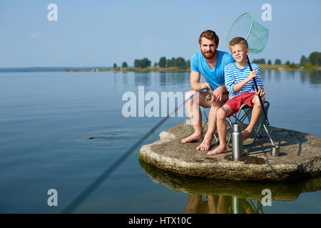 Portrait of handsome bearded father teaching his rascal teenage son to fish sitting on rock island in lake with fishing rods and gear on sunny summer  Stock Photo