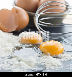 square image of two cracked eggs showing yellow yolk on a pile of white flour with a whisk and full eggs in the back ground, shot with focus of subjec Stock Photo