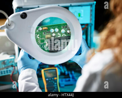 Engineer with white lab coat examining graphics card, looking through magnifying glasses to board, Austria Stock Photo