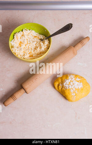 Fresh prepared yellow dough with white flour and wooden rolling pin on light marble background. Green bowl with cottage cheese or curd and dough for c Stock Photo
