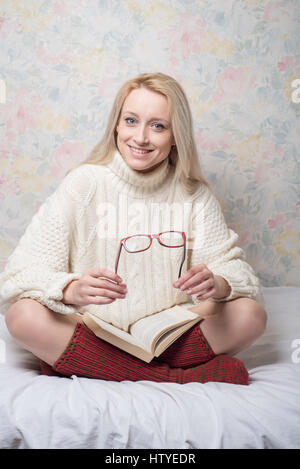 Woman sitting cross-legged on bed with book holding reading glasses Stock Photo