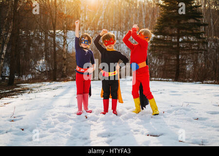 Three children standing on frozen lake wearing superhero costumes Stock Photo