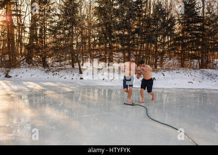 Two men in shorts running through sprinkler on frozen lake holding hands Stock Photo