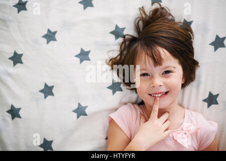 Overhead view of smiling girl lying in bed with finger on her lips Stock Photo