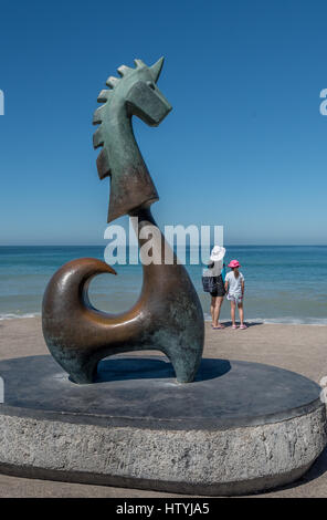 Mother and daughter stand on the Puerto Vallarta malecon looking at the blue sea from behind The Good Fortune Unicorn sculpture by Aníbal Riebeling. Stock Photo