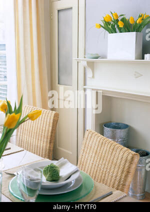 Wicker back chairs at table with white and green crockery in an economy style dining room with yellow striped curtains Stock Photo