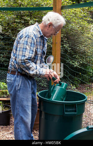 Man filling his watering can from a large garbage can of water beside his garden, in western Washington, USA Stock Photo