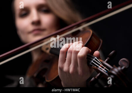 hand closeup detail over a blurred face of a young woman violinist player playing her instrument on her shoulder holding bow. portrait in a blurred da Stock Photo