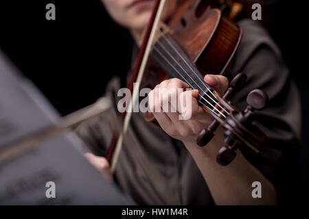 closeup of an unrecognizable musical player's hand holding a violin while playing it. Portrait on a black room with music score blurred in foreground Stock Photo