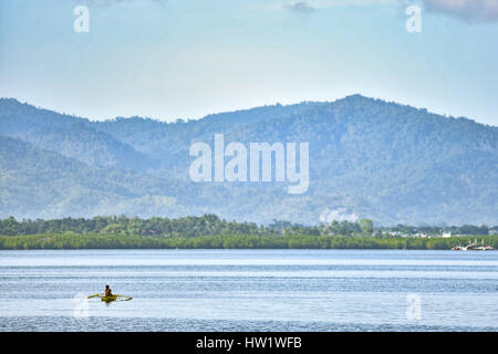 Alone Filipino fisherman in his traditional fishing boat out on the water with tree covered hills background. Stock Photo