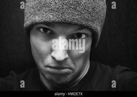 Young agressive Caucasian man in gray hat. Close-up studio face portrait over dark wooden wall background, selective focus, black and white photo Stock Photo