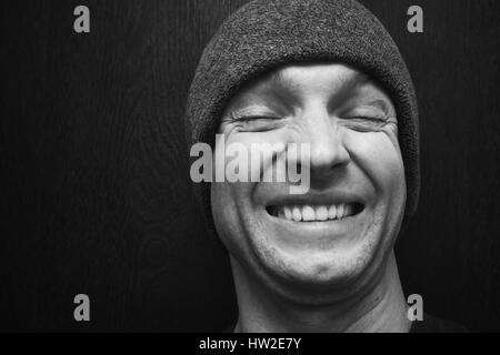 Young laughing man in gray hat. Closeup face portrait over dark wooden wall background, selective focus, black and white photo Stock Photo