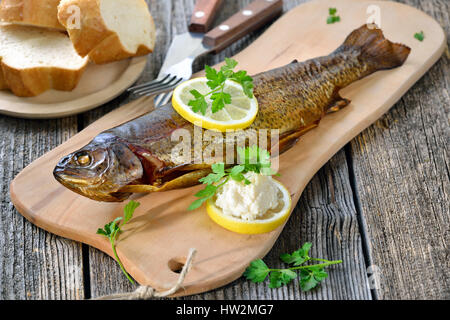 A smoked rainbow trout served on a wooden cutting board with horseradish, parsley, lemon  and baguette Stock Photo