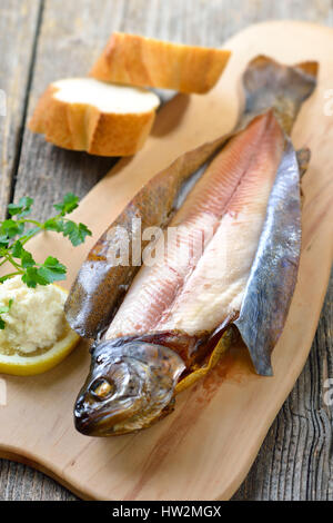 A smoked rainbow trout served on a wooden cutting board with horseradish, parsley, lemon  and baguette Stock Photo