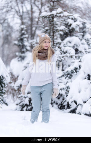 Attractive smiling young blonde girl walking in winter forest. Pretty woman in wintertime outdoor. Wearing winter clothes. Knitted sweater, scarf, hat Stock Photo