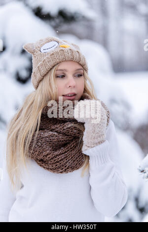 Attractive smiling young blonde girl walking in winter forest. Pretty woman in wintertime outdoor. Wearing winter clothes. Knitted sweater, scarf, hat Stock Photo