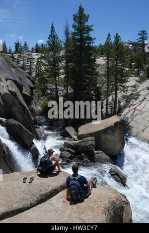 Two mid-adult men with backpacks on sit together and talk on a rock above a raging river. A pair of flip flops sits behind one of the men. Stock Photo