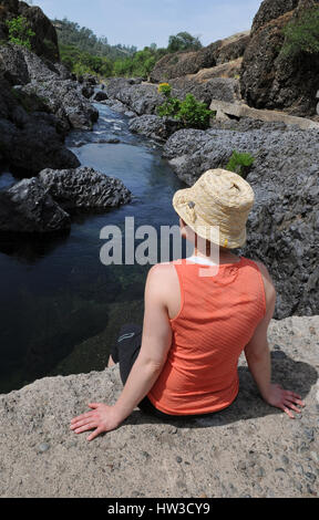 A mid-adult woman with her back to the camera sits on a rock above a river looking off to the side. She is wearing a peach tank top and a straw hat. Stock Photo