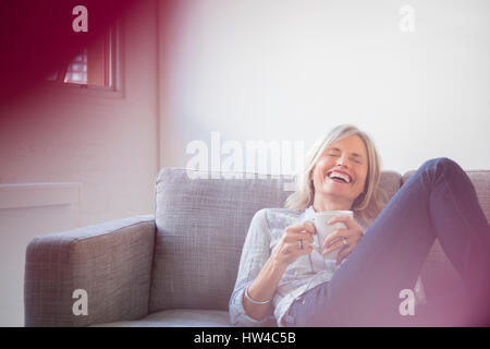 Laughing Caucasian woman sitting on sofa drinking coffee Stock Photo