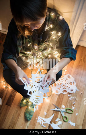 Mixed Race woman sitting on floor wrapped in string lights holding snowflake Stock Photo